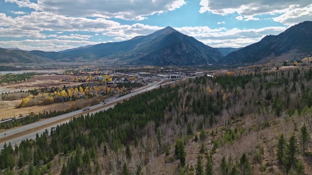 Movimento aéreo de câmera (dolly) inclinando para cima em direção a picos de montanhas acima de árvores perenes e desfolhadas, com carros trafegando na rodovia, em Frisco, Colorado.