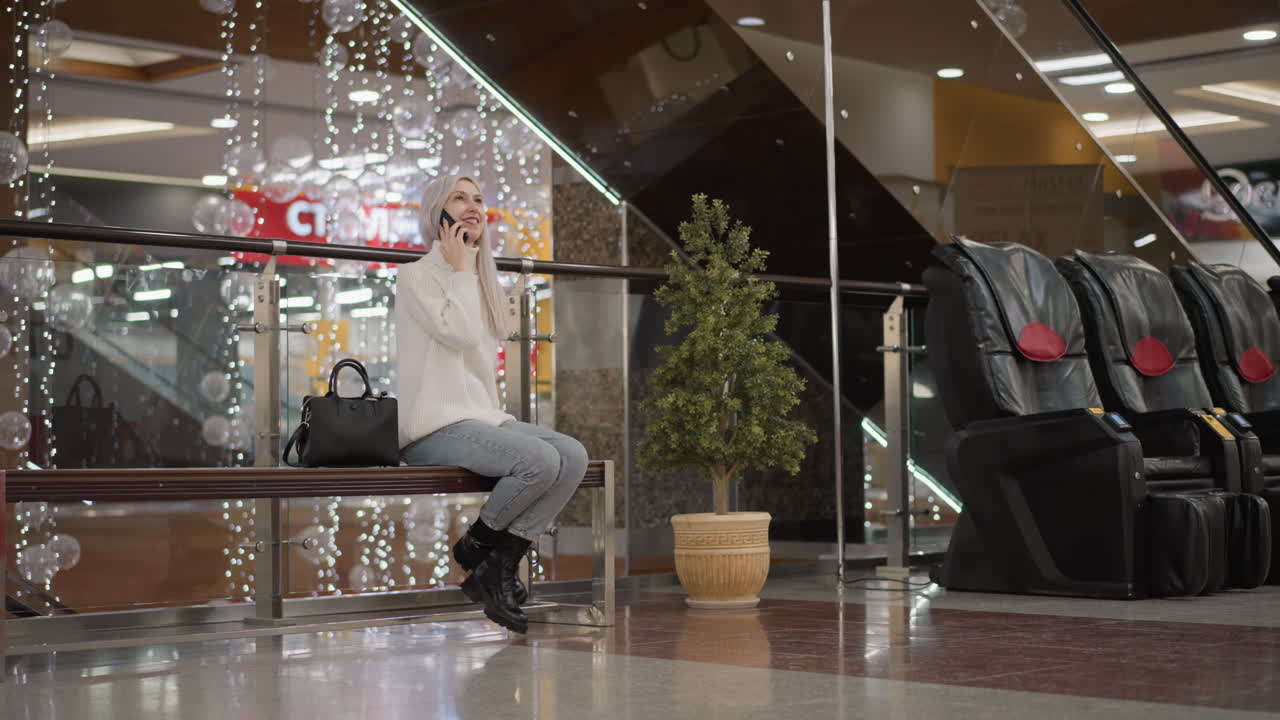 graceful woman seated on bench in mall lifts leg laughing into phone call, cozy white sweater with bag at side, modern interior backdrop with decorative lights escalator and office chairs nearby
