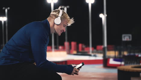 deportista sonriente sentado en el parque escuchando música con auriculares bluetooth, enviando mensajes de texto a su teléfono móvil y mirando a su alrededor mientras toma un descanso durante su sesión de entrenamiento por la noche 1