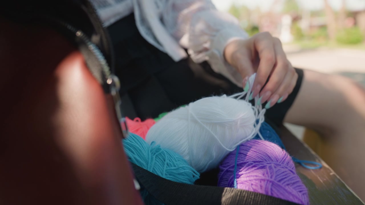 Primo piano di una giovane donna che seleziona il filato da gomitoli colorati in un ambiente esterno. La sua mano con unghie dipinte interagisce delicatamente scegliendo matasse bianche e viola tra tonalità blu, rosa e arancioni. Morbido.