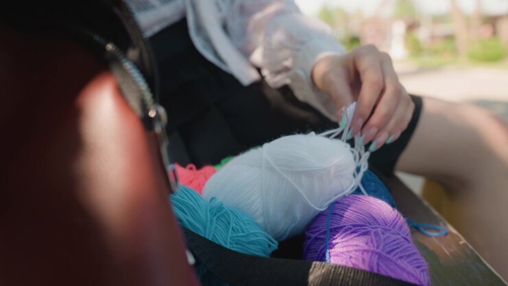 Close up of young woman selecting yarn from colorful balls in outdoor setting. Her hand with painted nails interacts gently choosing white and purple skeins among blue pink orange hues. Soft