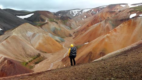 Aerial, rising, drone shot over a hiker standing on a hill, looking down on a valley, in Laudmannalauger, Iceland