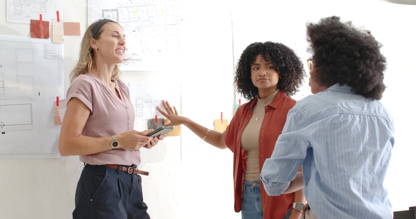 Diverse female team moving to studio wall after colleague gesturing with tablet, reviewing swatches
