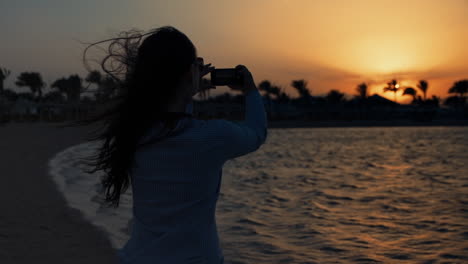 mujer atractiva tomando una foto del atardecer mar en el teléfono móvil en la orilla del mar.