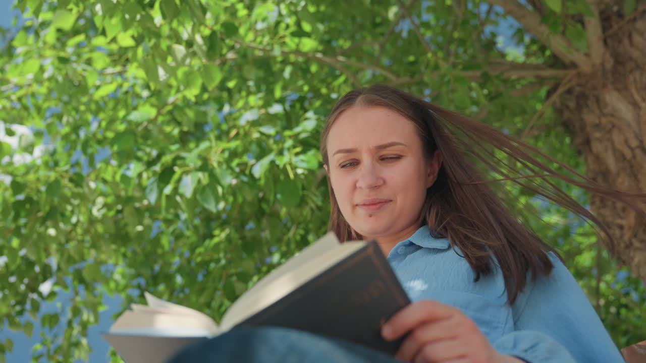 Mujer leyendo al aire libre, señora disfrutando de un libro bajo los árboles, mujer relajada leyendo un libro bajo el fresco dosel de un árbol frondoso, mujer serena absorta en la lectura de un cuento bajo la fresca sombra de un gran árbol