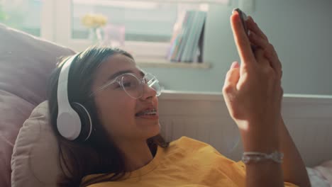 caucasian teenage girl browsing phone with smile and wearing headphones while lying on bed