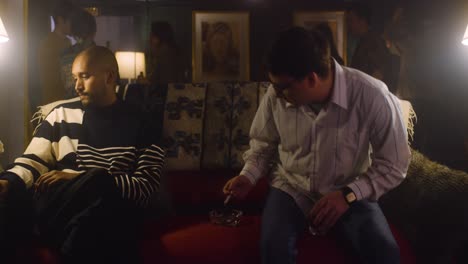 two men talk on couch at house party.