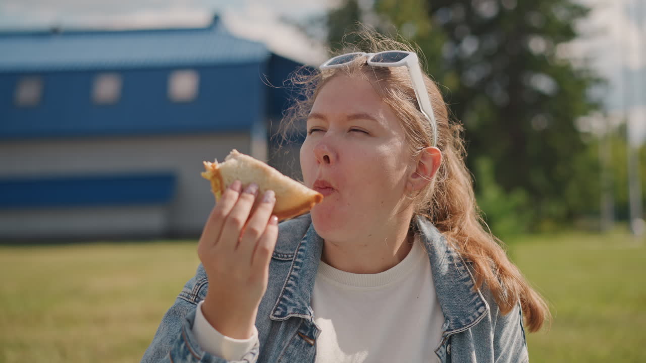 Junge weiße Frau isst Sandwich im Freien während eines sonnigen Picknicks im Vorort, Jeansjacke und Sonnenbrille, spielerischer Bissen und Krümel, ehrliche Nahaufnahme, die einen freudigen Ausdruck einfängt, Studentin und reisende Feinschmeckerin
