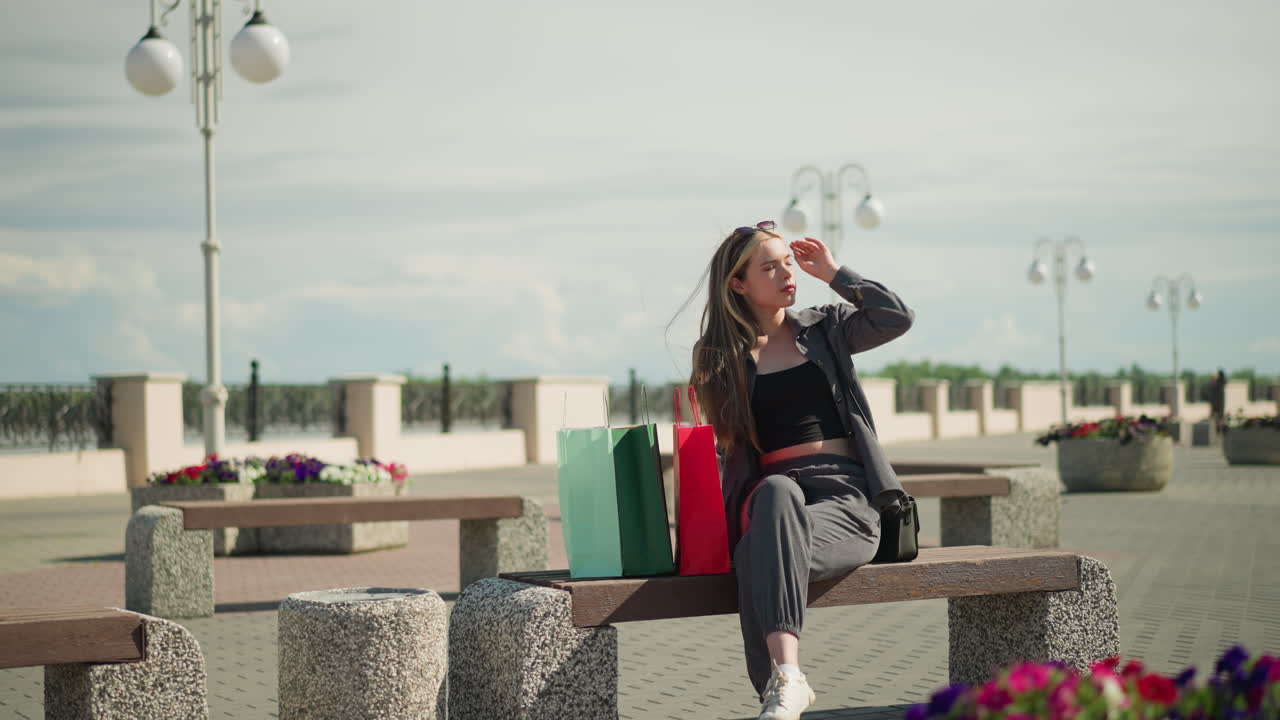 그녀의 에 세 개의 다채로운 쇼핑 가방을 가진 야외 벤치에 앉아있는 여성, 그녀는 다리를 고 머리를 조정하고, 은 날에 세련된 포즈로 위를 바라보고, 주위에 전기 기둥을 가지고 있습니다.