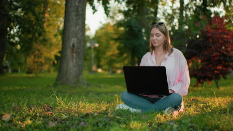 lady looks up thoughtfully with a warm smile as she continues working on her laptop while seated cross-legged on a grassy field, background features trees and greenery with natural lighting