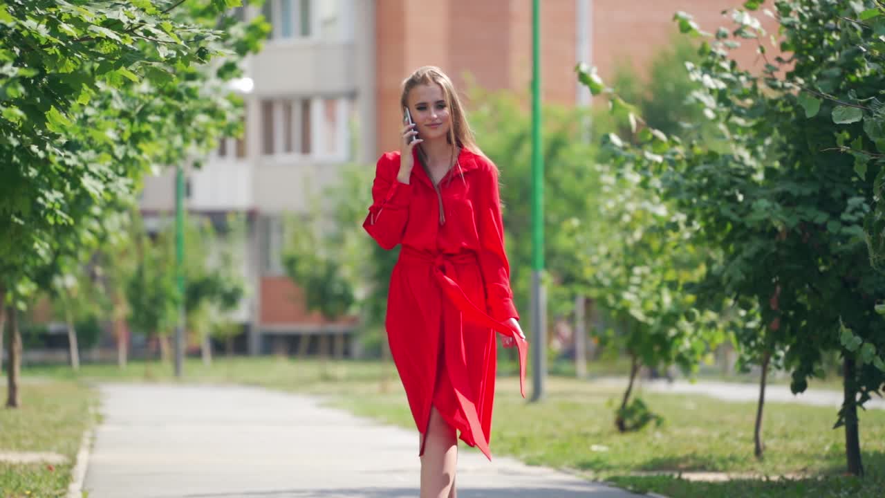 Sexy woman in red dress outdoors. Young lady walking slowly on the street and talking on the mobile phone. Beautiful female in passionate dress in a summertime.