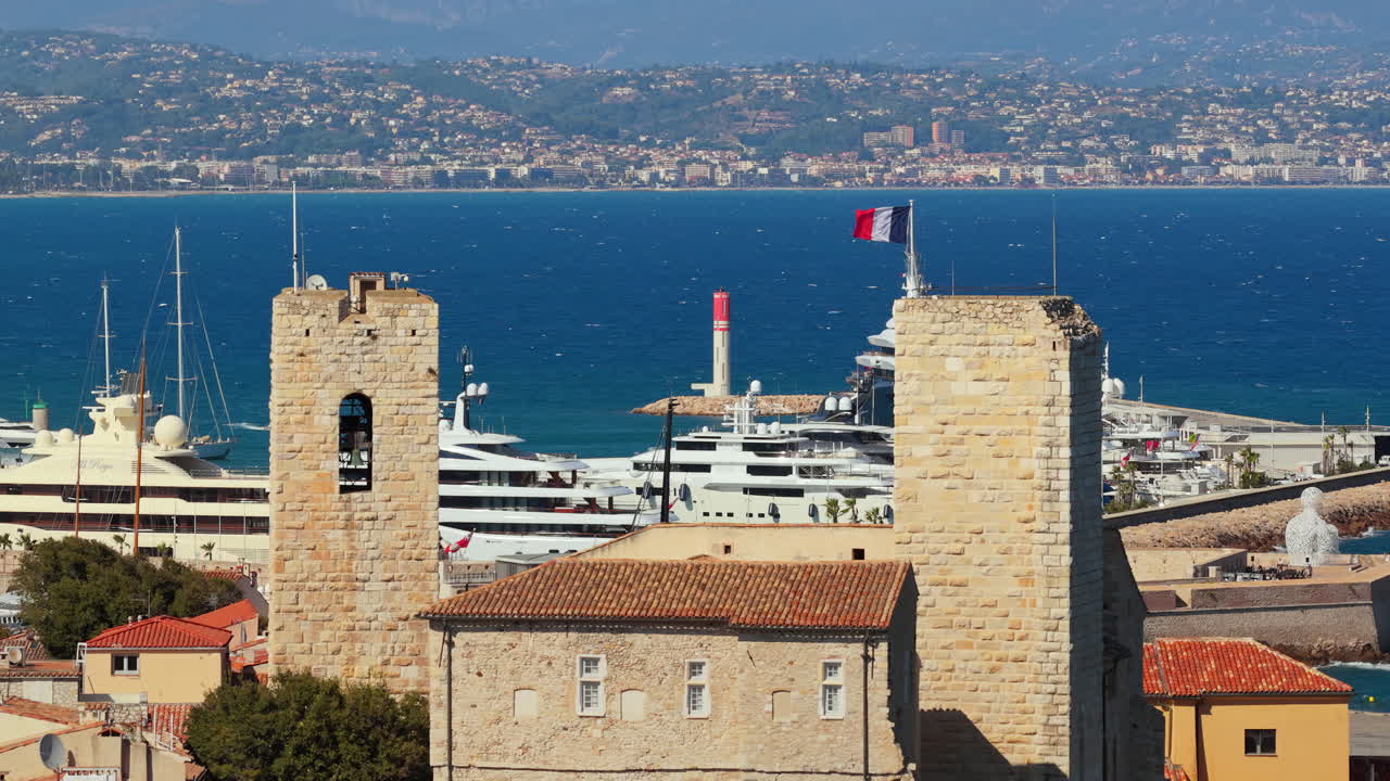 Aerial drone view of terracotta rooftops in Antibes Old Town with the medieval bell tower of Chateau Grimaldi, luxury yachts docked in the marina behind