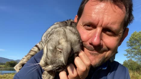 Close-up-of-caucasian-man-strocks-cute-cat-hanging-on-shoulder-outside-in-nature