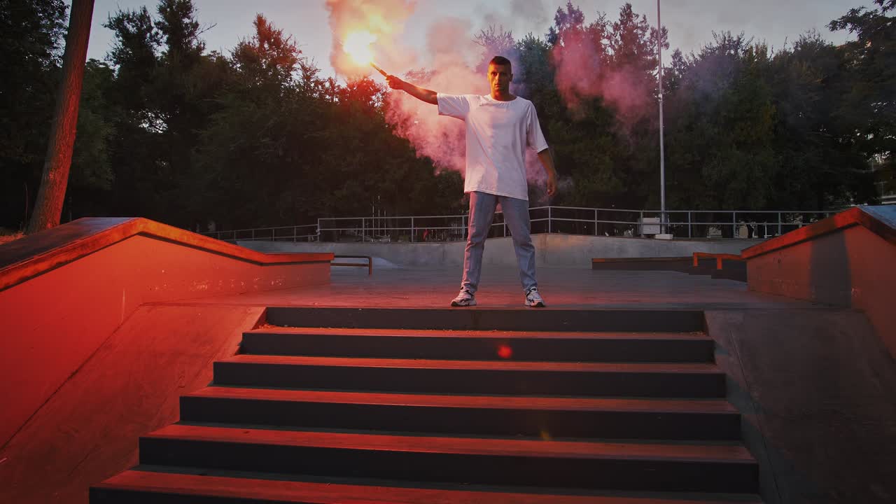 Un giovane tiene in mano un fumogeno rosso luminoso e si prepara a eseguire breakdance in piedi sulle scale di un rollerdrome di sera. Movimento lento.