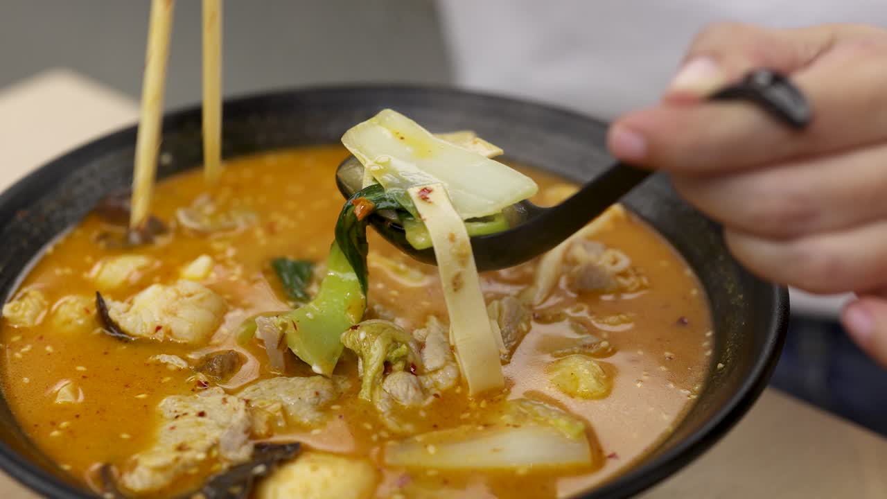 Close-up of chopsticks and spoon lifting ingredients from steaming spicy hotpot under soft lighting