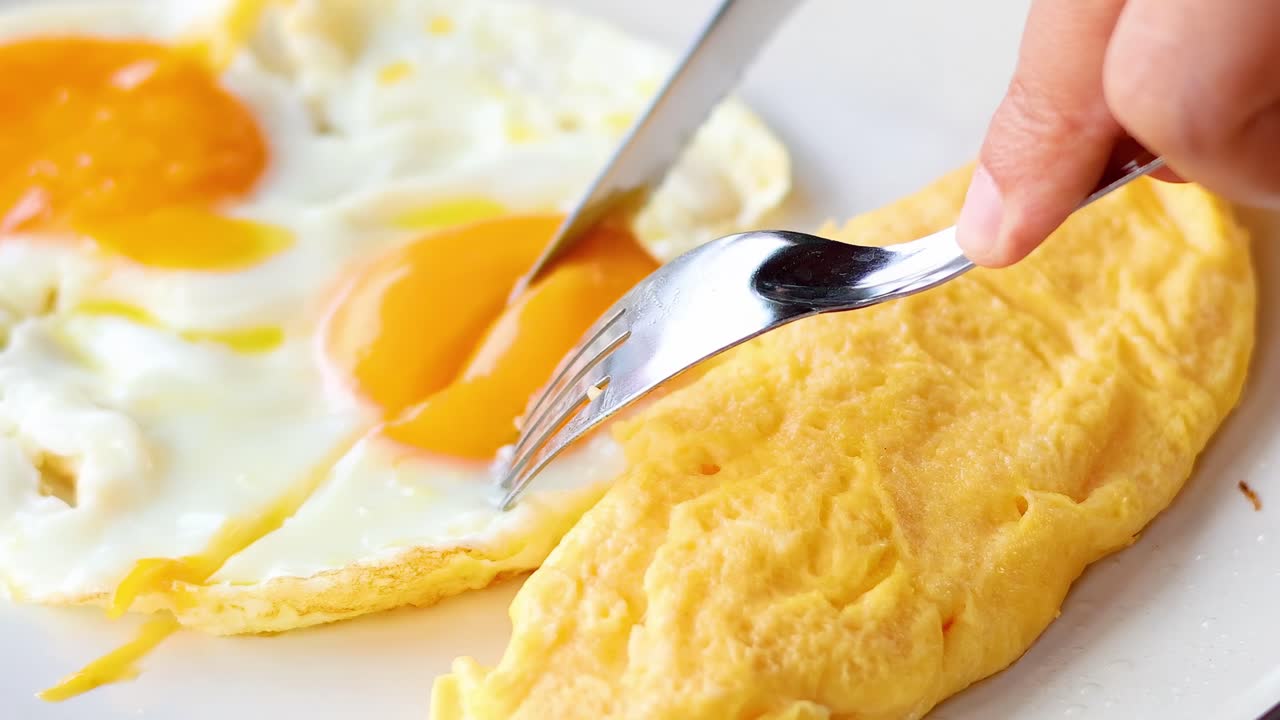 Close-up of a knife and fork cutting into an omelet and sunny-side up eggs on a plate.