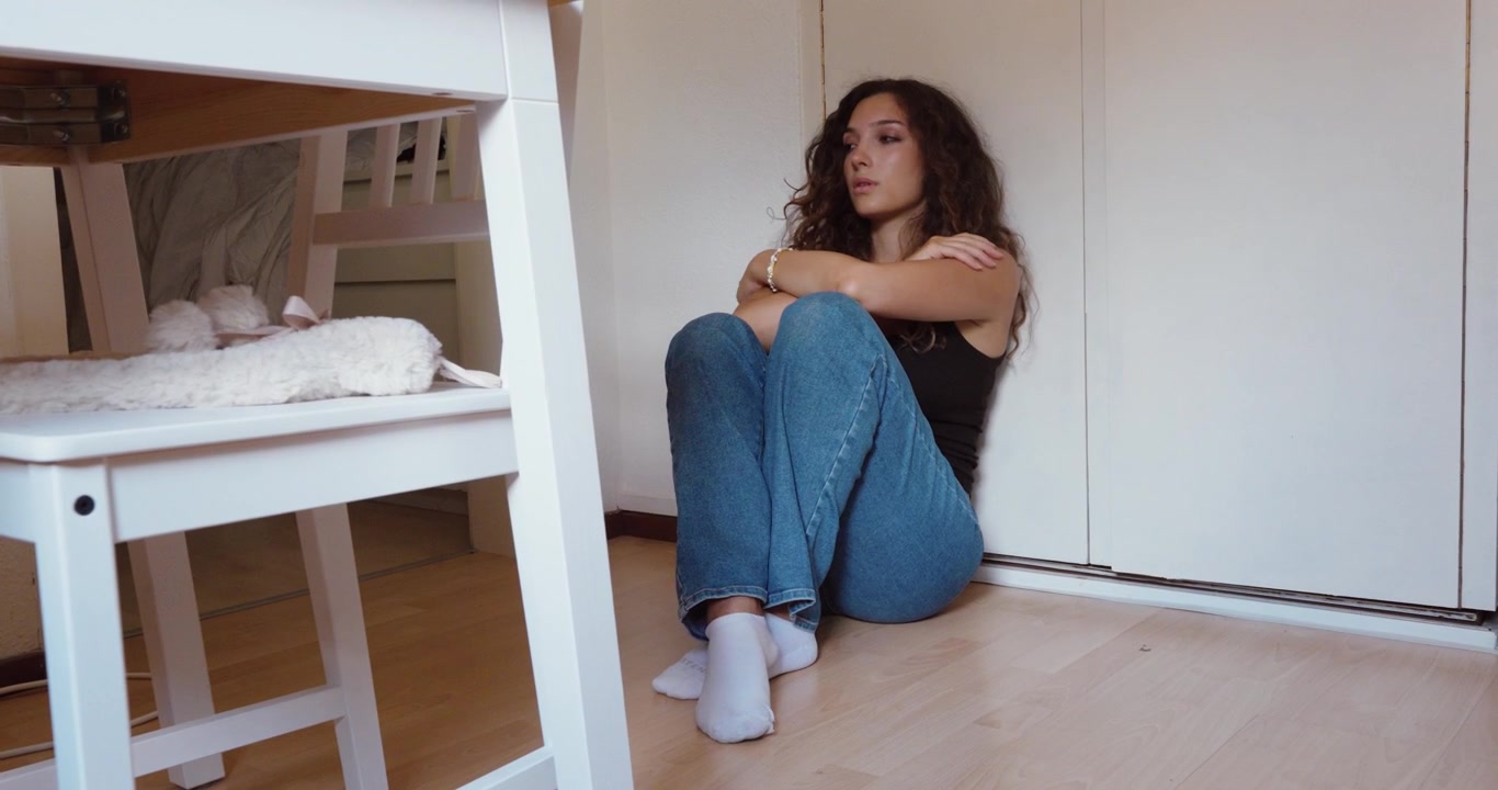 A sad and lonely woman sits on the floor while experiencing emotional distress and feelings of isolation at home.