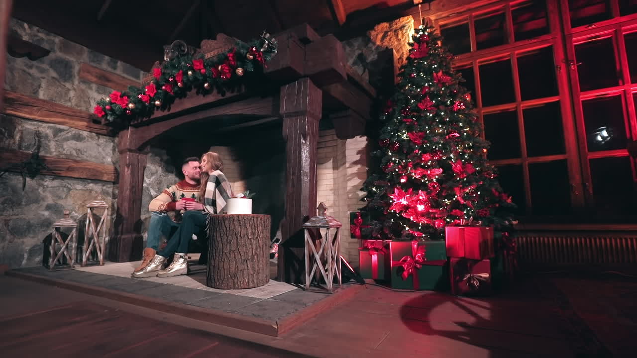 Young people in love near fireplace at Christmas. Happy man and woman sitting together in the country house. Beautiful Christmas tree in the room.