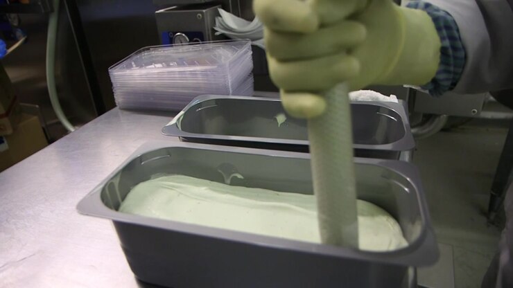 A worker wearing a protective glove filling a stainless steel tray with fresh, creamy gelato in an industrial ice cream laboratory setting.