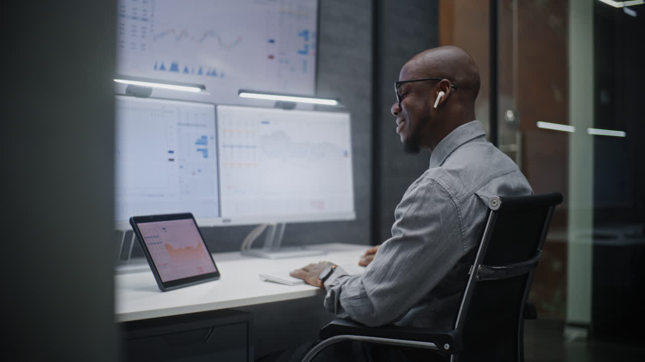 Financial Analyst Talking in Wireless Headphones, Monitoring Exchange Market Charts on Computer with Multi-Monitor Workstation. African American Businessman Works in Office at Night. Vertical Shot.