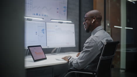 financial analyst talking in wireless headphones, monitoring exchange market charts on computer with multi-monitor workstation. african american businessman works in office at night. vertical shot.