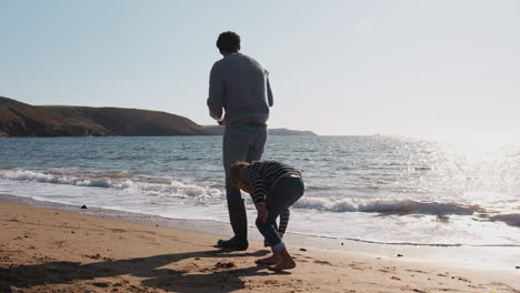 father and son walking along beach by breaking waves on beach collecting stones to skim