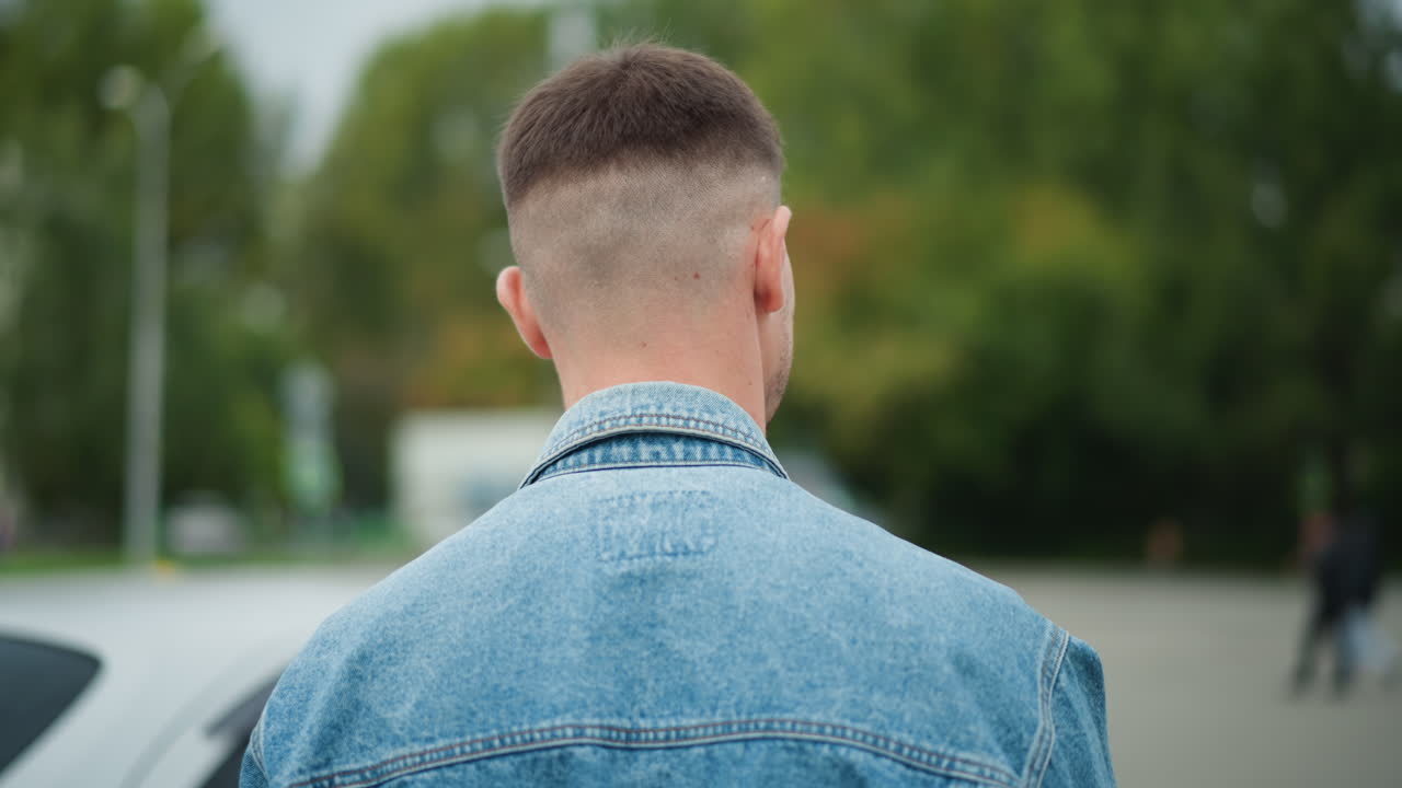 man back portrait facing empty lot, short haircut and denim jacket, slow turn to open car door and enter, closeup shoulder and head shot, intimate anticipation and quiet suburban greenery