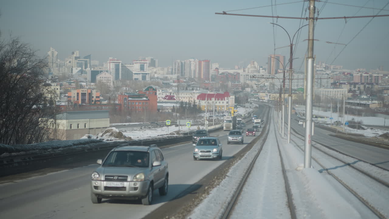 View from moving train on center tracks between car lanes shows winter city skyline, traffic flowing along road, snow lined rails, distant buildings under pale sky, urban transit perspective