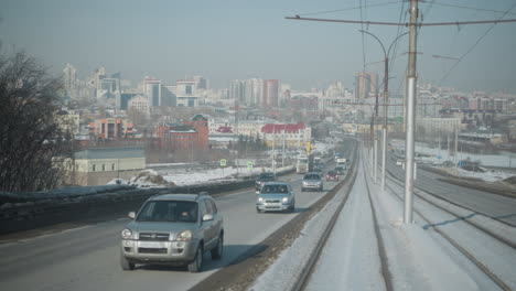 view from moving train on center tracks between car lanes shows winter city skyline, traffic flowing along road, snow lined rails, distant buildings under pale sky, urban transit perspective