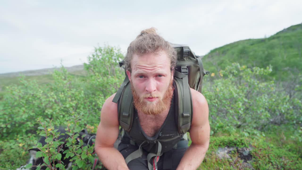 A Bearded Male Hiker Looking Closely To The Camera While Sitting Beside His Dog In Grassy Field At Anderdalen National Park In Senja, Norway
