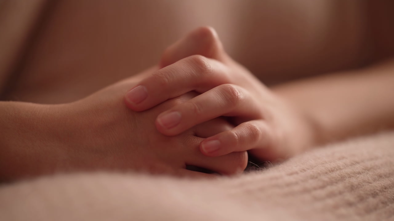Close Up Caucasian Hands Clasped Gently On Textured Blanket In Warm Low Light, Intimate Spa Bedroom Mood,