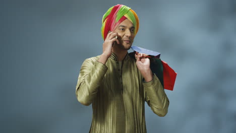 young cheerful indian man in traditional clothes and turban talking on the phone and holding shopping bags