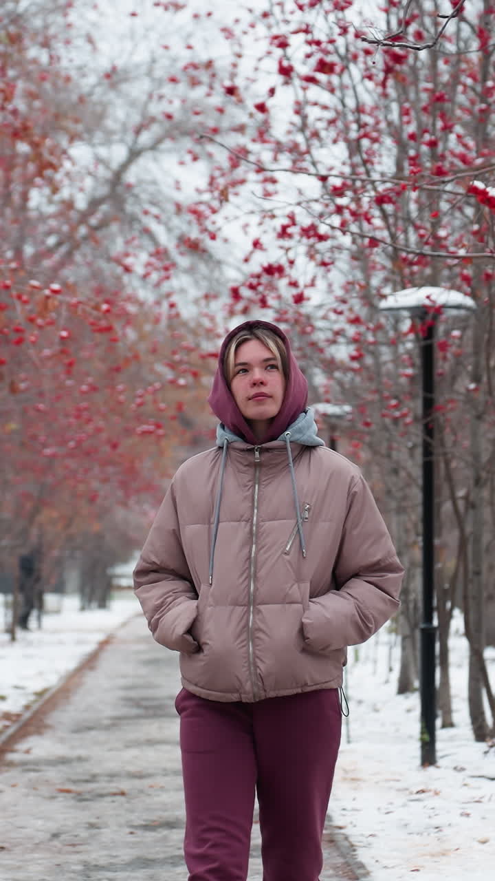 joven con chaqueta cálida y capucha camina por un camino nevado bordeado de árboles de bayas rojas, mirando a su alrededor con curiosidad, distante figura borrosa en el fondo