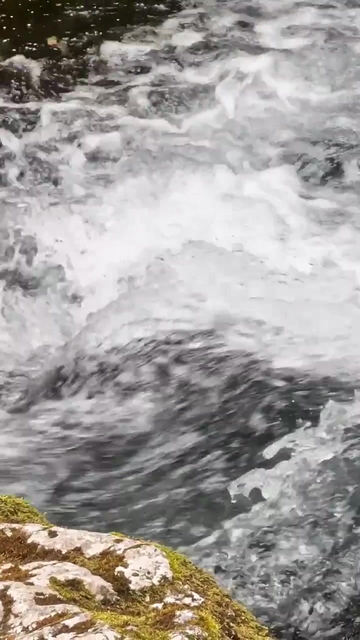 Primo piano del deflusso spumeggiante della cascata di Golling in Austria, con acqua bianca impetuosa che scorre su rocce scure del ruscello e un bordo di pietra muschioso.