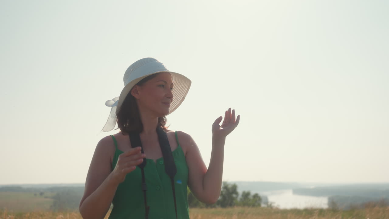 woman in green dress puts on wide hat while gently tucking hair back behind shoulders in open field, camera hanging from neck, bright summer light and distant river landscape