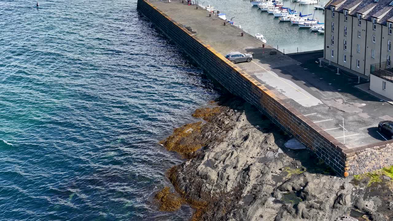 Drone glides above Fife harbor, revealing marina, rocky shoreline, historic buildings, and parked cars