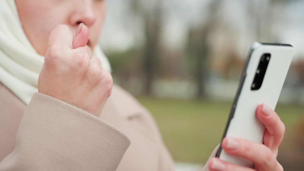 Donna che tiene lo smartphone all'aperto controllando labbra e messaggi, primo piano delle mani con cappotto beige e sciarpa bianca, sottile gesto ansioso, sfondo di parco nuvoloso, unghie curate che toccano il touchscreen, moderno.