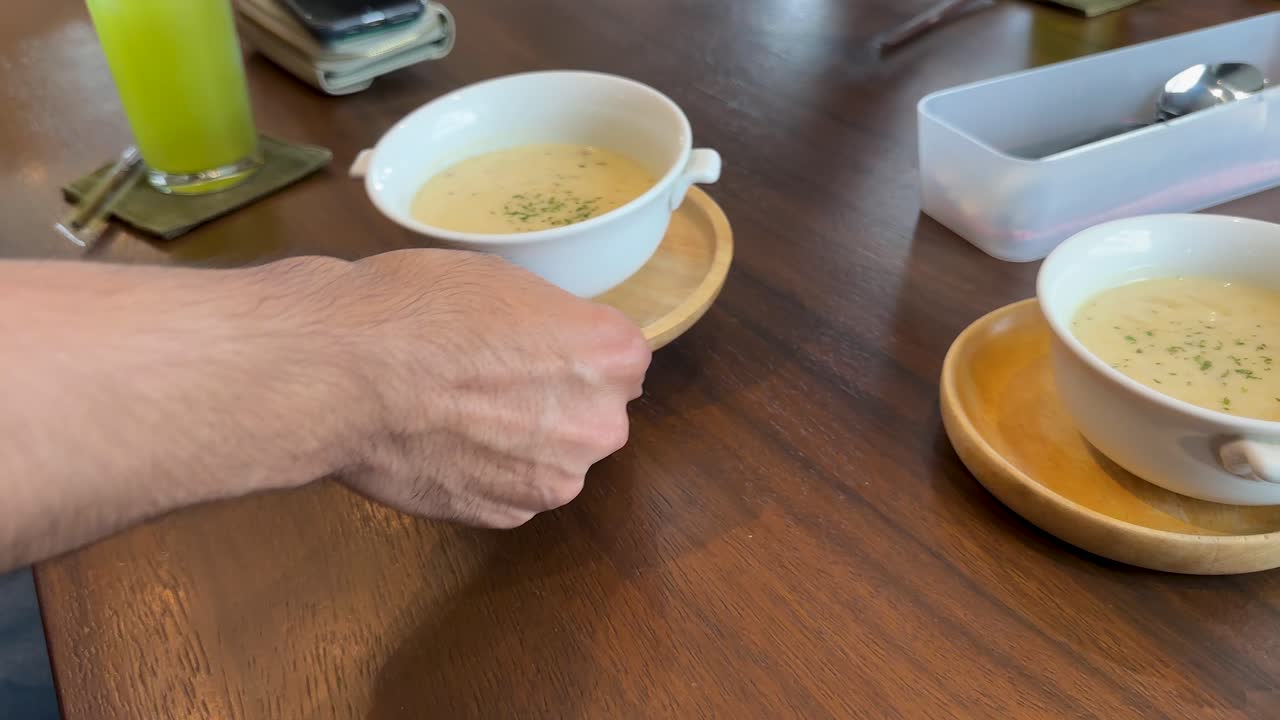 Hand sets down cream soup bowl on wooden table in cozy, naturally lit Bangkok cafe