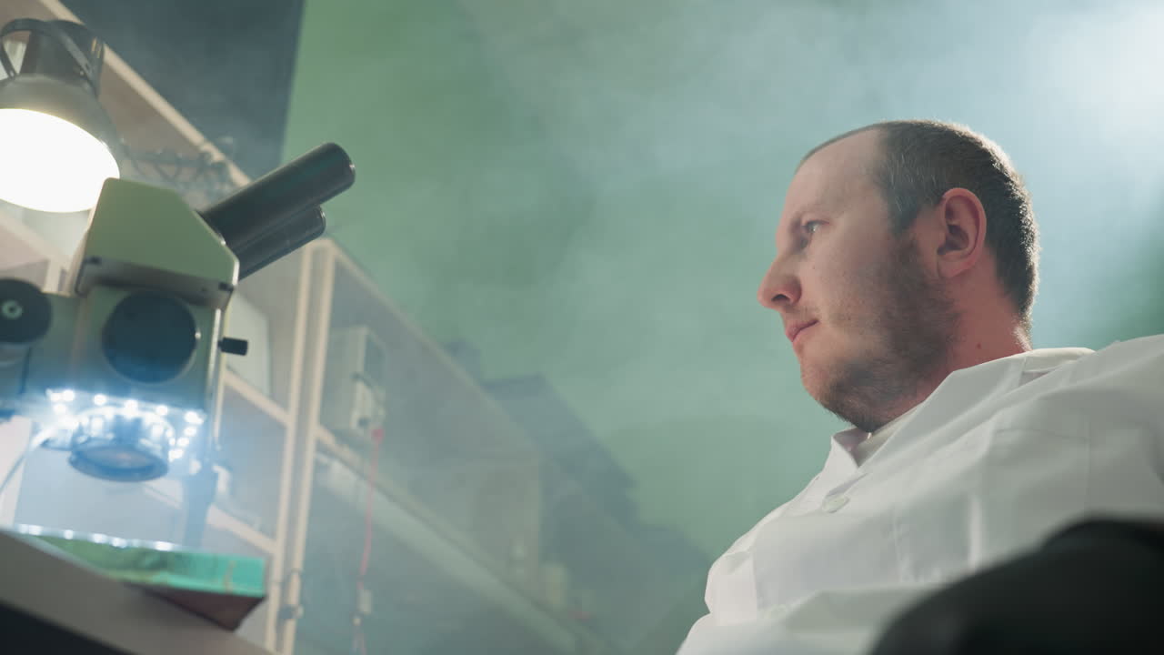una vista de cerca de un científico en una bata de laboratorio blanca, sentado y descansando con los ojos cerrados, la mano en la frente, en un entorno de laboratorio, con estantes y un microscopio encendido y reflejo de luz