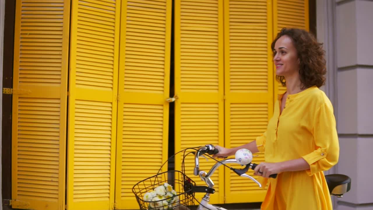Smiling young woman in a long yellow dress walking by the yellow wooden fence on the window holding her city bicycle handlebar