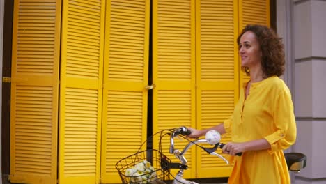 Smiling-young-woman-in-a-long-yellow-dress-walking-by-the-yellow-wooden-fence-on-the-window-holding-her-city-bicycle-handlebar