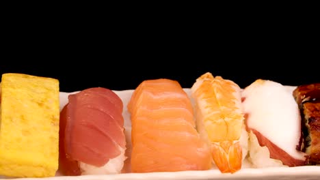 a variety of sushi nigiri and sashimi, including tamago, tuna, salmon, shrimp, squid, eel, and salmon roe, rotates on a white plate against a black background under bright studio lighting