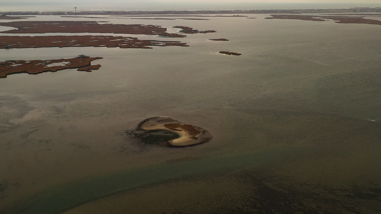 A top-down aerial view over a salt marsh, it is low tide - you can see a sandbar in the water. The camera dolly in - tilts down towards the sand mound in the dark green waters.