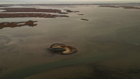 A top-down aerial view over a salt marsh, it is low tide - you can see a sandbar in the water. The camera dolly in - tilts down towards the sand mound in the dark green waters.