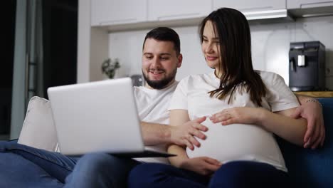 una hermosa mujer embarazada y un hombre navegan por la red sentados en el sofá con una computadora portátil