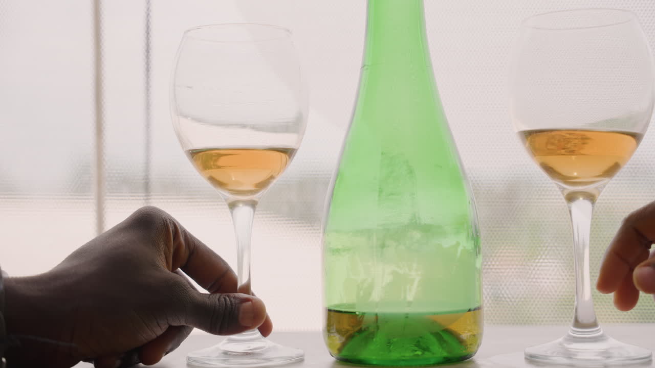 Romantisk vinaften med sort par, der deler ravfarvet vin fra grøn flaske og stilkglas ved et gennemsigtigt vindue, hænder rækker ud for at skåle over solbeskinnet bord, kondens på flasken, intim hyggelig stemning