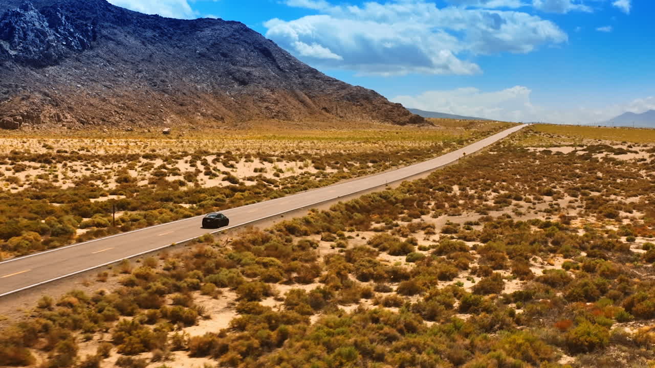 Black car driving by the road in the desert of Nevada. Beautiful mountains at backdrop with no vegetation on. Aerial view.