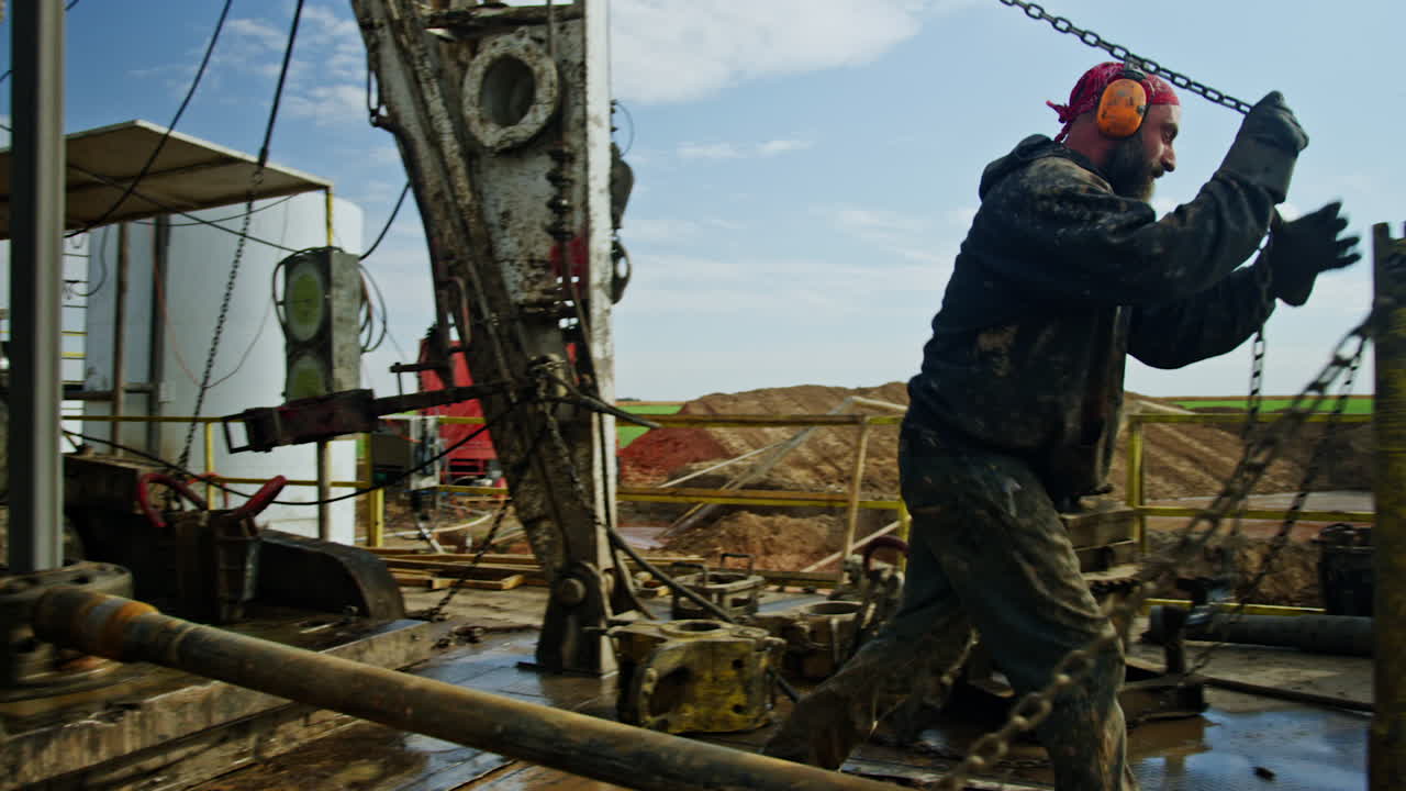 Man wearing dirty clothes is pulling the pipe hanging on the chains. Worker goes to the next pipe in a pile to attach another to the chain.