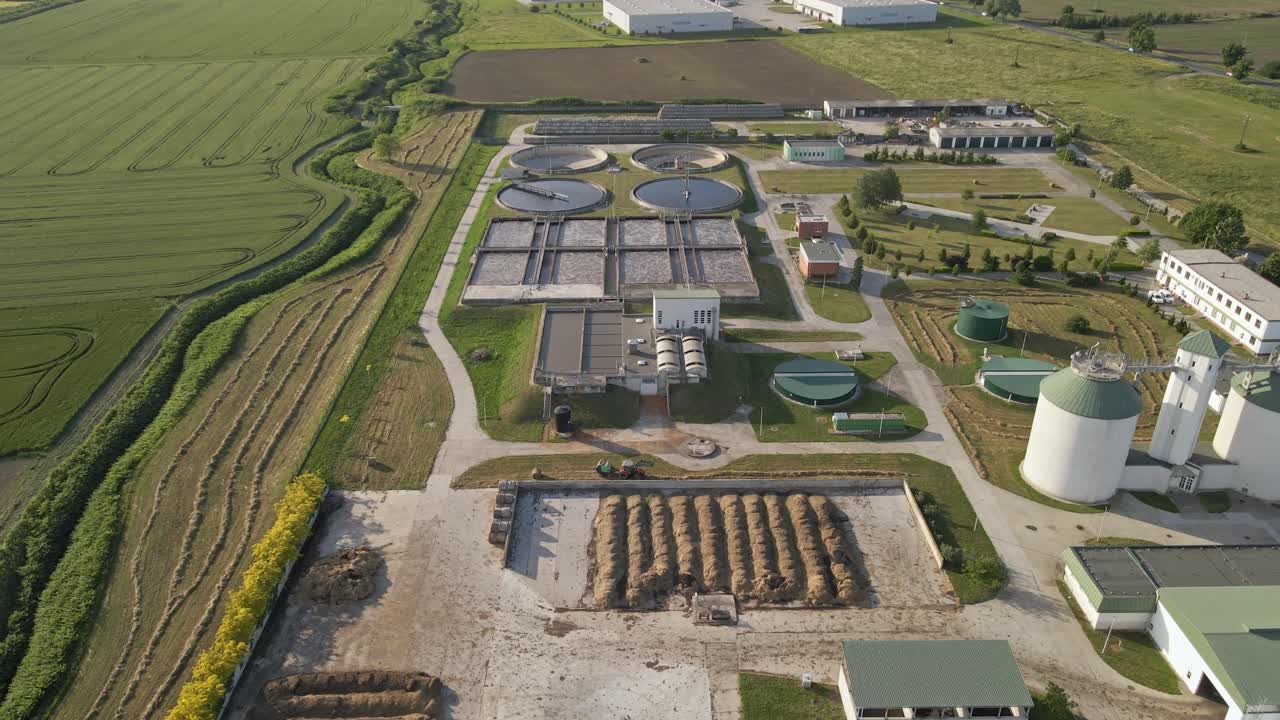 An aerial video of a modern wastewater treatment facility surrounded by green fields. The plant features multiple circular and rectangular treatment tanks and storage silos.