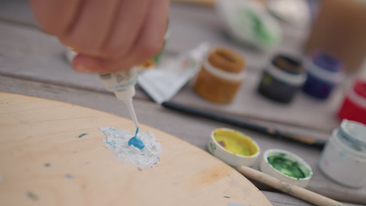 close de uma mão misturando tinta azul em uma paleta, ambiente de rua ao ar livre com pequenos potes e tubos, mesa de madeira, artista preparando pigmentos, luz suave do dia, foco seletivo, textura tátil se formando, sketch